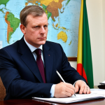 A professional British diplomat, fully clothed in a modest, dark business suit, seated at a polished mahogany table in a stately, well-appointed office. Subtle, strategic maps of the Niger Delta region are visible in the blurred background, hinting at economic interests. The diplomat holds a pen, poised to write, with a thoughtful and focused expression. Natural pose, perfect anatomy, correct proportions, well-formed hands, proper finger count, natural body proportions, safe for work, appropriate content, fully clothed, professional, modest, family-friendly, high-quality professional photography.